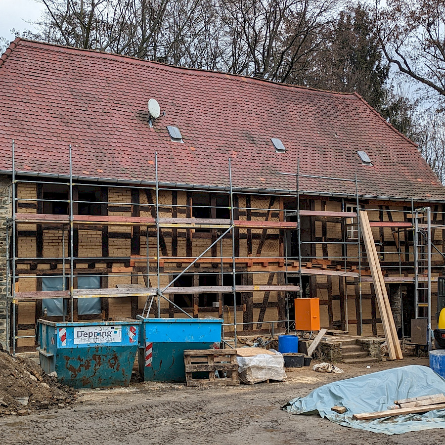 Ansicht des Haupthauses mit Baugerüst, davor Container, Baumaterialen und ein gelber Bagger