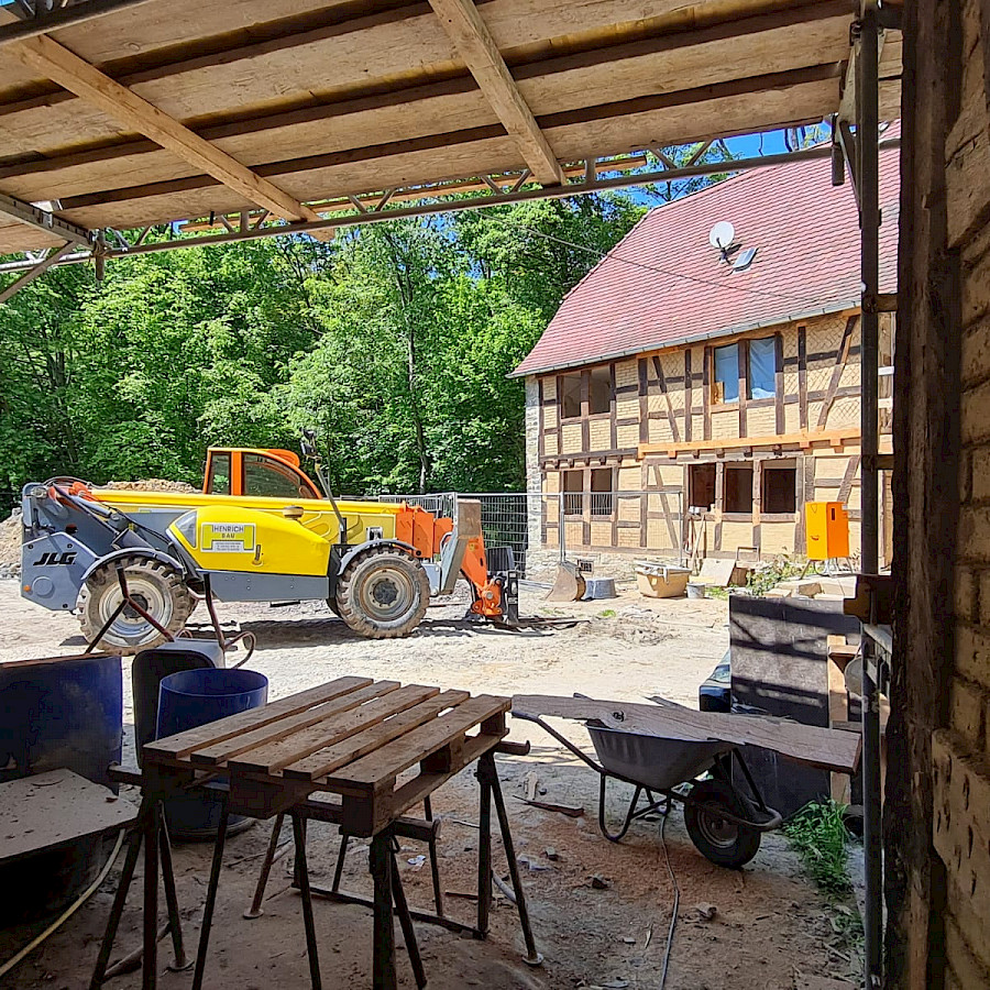 Blick aus dem Torhaus, in dem Arbeitsmaterialien stehen, auf den Hof in dem einer Baufahrzeug steht, dahinter das Haupthaus