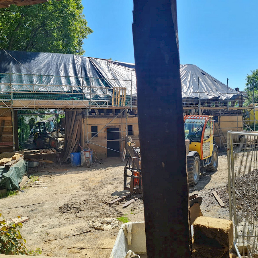 Scheunenhaus mit Gerüst und Plane über dem freigelegtem Dachstuhl, davor ein Bagger und ein Bauzaun, Blick vom Hauptgebäude