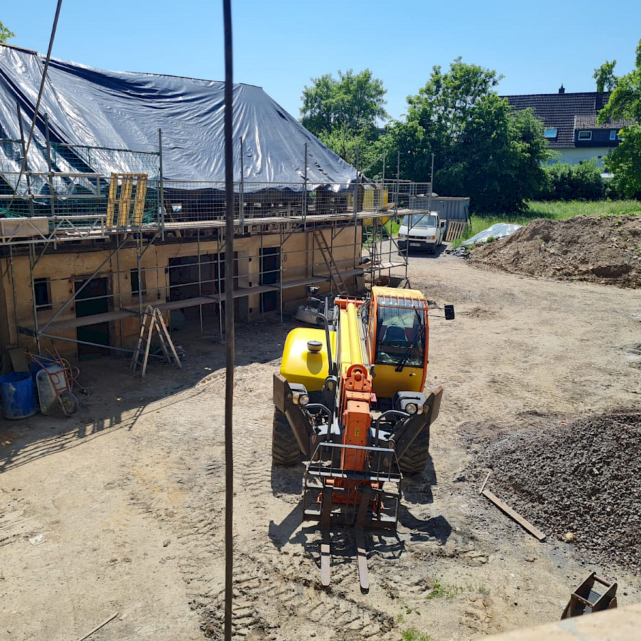 Scheunenhaus mit Gerüst und Plane über dem freigelegtem Dachstuhl, davor ein Bagger, Blick vom Hauptgebäude