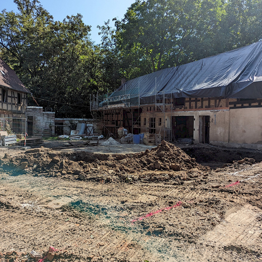 Blick über die ausgehobene Baugrube auf des Scheunenhaus, dessen Dachstuhl mit einer Plane abgedeckt ist