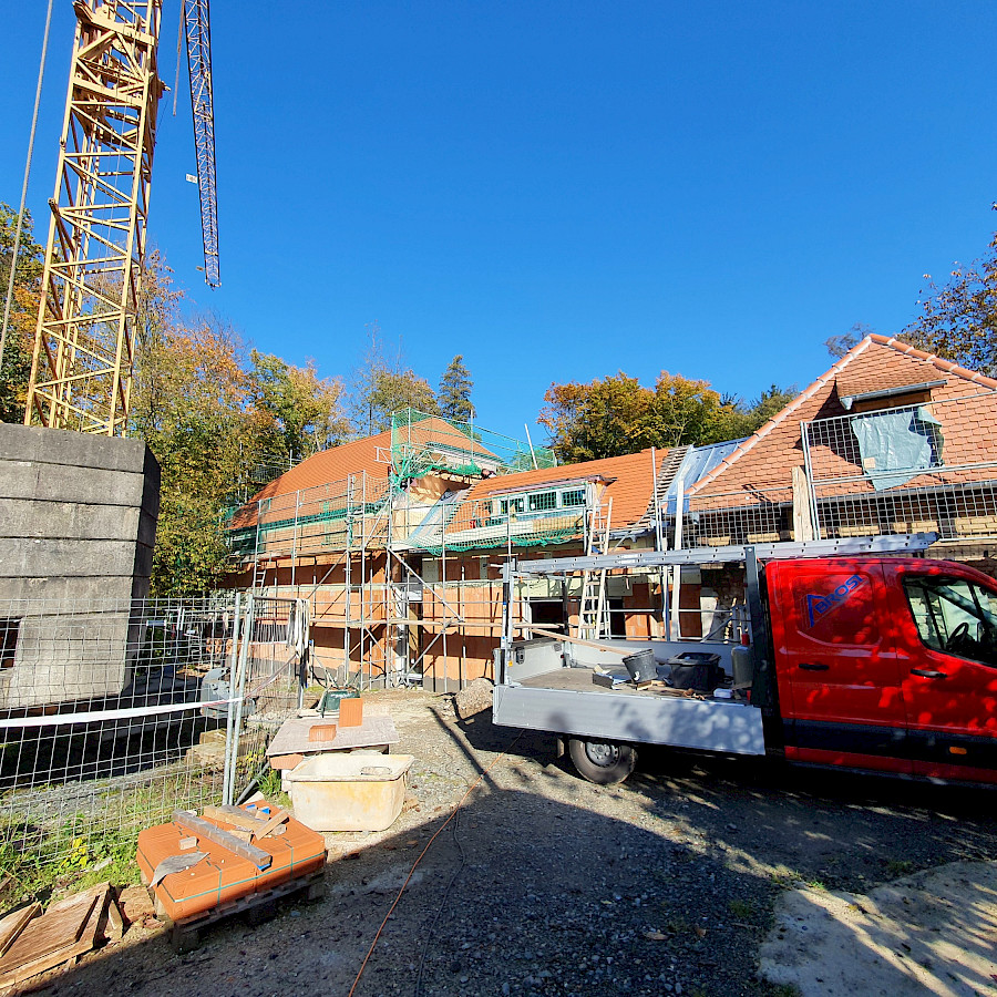 Blick von der Streuobstwiese auf die fertig gemauerten und gedeckten Zwischenbauten, davor ein Baukran und ein Lieferwagen
